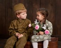 Children boy are dressed as soldier in retro military uniforms and girl in pink dress sitting on old suitcase, dark wood backgroun Royalty Free Stock Photo