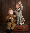 Children boy are dressed as soldier in retro military uniforms and girl in pink dress sitting on old suitcase, dark wood backgroun Royalty Free Stock Photo