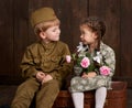 Children boy are dressed as soldier in retro military uniforms and girl in pink dress sitting on old suitcase, dark wood backgroun Royalty Free Stock Photo