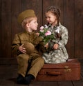 Children boy are dressed as soldier in retro military uniforms and girl in pink dress sitting on old suitcase, dark wood backgroun Royalty Free Stock Photo