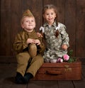 Children boy are dressed as soldier in retro military uniforms and girl in pink dress sitting on old suitcase, dark wood backgroun Royalty Free Stock Photo