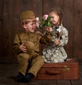 Children boy are dressed as soldier in retro military uniforms and girl in pink dress sitting on old suitcase, dark wood backgroun Royalty Free Stock Photo