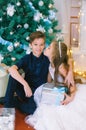 Children, a boy and a girl in a white ball gown near a Christmas tree in holiday dresses with gifts and silver confetti.