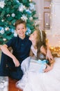 Children, a boy and a girl in a white ball gown near a Christmas tree in holiday dresses with gifts and silver confetti.