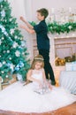 Children, a boy and a girl in a white ball gown near a Christmas tree in holiday dresses with gifts and silver confetti. Royalty Free Stock Photo