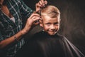 Children hairdresser with scissors is cutting little boy against a dark background. Contented cute preschooler boy