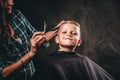 Children hairdresser with scissors is cutting little boy against a dark background. Contented cute preschooler boy