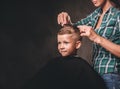 Children hairdresser with scissors is cutting little boy against a dark background. Contented cute preschooler boy