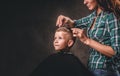 Children hairdresser with scissors is cutting little boy against a dark background. Contented cute preschooler boy