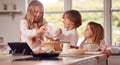 Children In Pyjamas Making Pancakes In Kitchen At Home Following Recipe On Digital Tablet