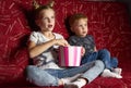 Children`s cinema: A girl and a boy watch a movie at home on a big red sofa in the dark and eat popcorn. Royalty Free Stock Photo