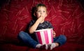 Children`s cinema: A little boy, a preschooler, watch a movie at home on a big red sofa and eat popcorn from a big Royalty Free Stock Photo