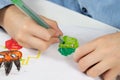 Children`s hands hold a green felt-tip pen and draw a green guy on a white piece of paper.