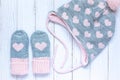 Children`s winter accessories, warm knitted mittens and hat - gray with pink hearts on white wooden background.