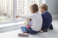 children sit on the floor by the window and admire the view. little blond boy and girl with pigtails pointing at something outside