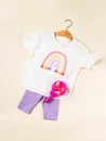 A children's T-shirt with a rainbow on a hanger and lilac shorts on a beige background. Children's clothing for