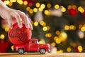 A childs hand puts a Christmas ball in the back of a red retro pickup truck against the backdrop of Christmas tree lights. Merry