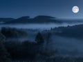 Chilling scenery of forested hills in fog with the full moon in the blue sky at night time Royalty Free Stock Photo