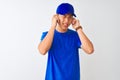 Chinese deliveryman wearing blue t-shirt and cap standing over isolated white background covering ears with fingers with annoyed
