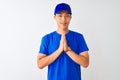 Chinese deliveryman wearing blue t-shirt and cap standing over isolated white background praying with hands together asking for