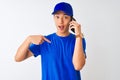 Chinese deliveryman wearing cap talking on the smartphone over isolated white background with surprise face pointing finger to
