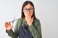 Chinese hairdresser woman wearing glasses holding scissors over isolated white background cover mouth with hand shocked with shame Royalty Free Stock Photo