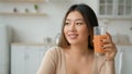 Chinese Korean woman drinking peach orange juice at home kitchen dreaming Asian girl drink glass of fruity smoothie low Royalty Free Stock Photo