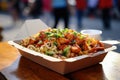 Chinese noodles with chicken and vegetables in cardboard box on table in street eatery, asian fast food, concept fast take-away