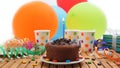 Chocolate birthday cake with a blue candle burning on rustic wooden table with background of colorful balloons, gifts Royalty Free Stock Photo