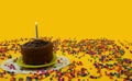 Chocolate birthday cake with 1 blue and white candle lit, on a small green plate, surrounded by candy balls scattered on a yellow Royalty Free Stock Photo