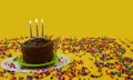 Chocolate birthday cake with 3 blue and white candles lit, on a small green plate, surrounded by candy balls scattered on a yellow Royalty Free Stock Photo