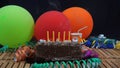 Chocolate birthday cake with five yellow candles extinguished on rustic wooden table with background of colorful balloons Royalty Free Stock Photo