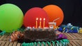 Chocolate birthday cake with three yellow candles burning on rustic wooden table with background of colorful balloons Royalty Free Stock Photo