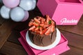 Chocolate cake with strawberries, basil leaves and chocolate plates around, on a pink box top view. Balloons in the background.