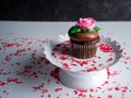 Chocolate cupcake with chocolate frosting and a pink rose on a white raised platter with red and pink sprinkles scattered like