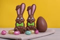 Chocolate Easter Bunnies And Eggs On White Wooden Table Against Yellow Background