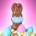 Chocolate Easter bunny, surrounded by Easter eggs, wearing a medical face mask for protection to the coronavirus