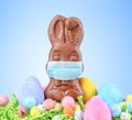 Chocolate Easter bunny, surrounded by Easter eggs, wearing a medical face mask for protection to the coronavirus