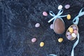 chocolate egg with blue bunny ear bows and multicolored chocolate eggs on a black textured background. Royalty Free Stock Photo