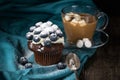 Chocolate muffin with soft cream and fresh blueberry powdered with sugar, cup of coffee with marshmallows, rustic background