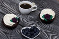 Chocolate muffins with cream flower. A cup of black coffee. Saucer with blueberries. Lies on pine boards painted black and white