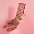 Chocolate pink bar holds a female hand on a pink background burst paper hole. Food concept art. Square frame Royalty Free Stock Photo