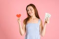 The choice between love and money. A woman holds money and a red heart symbol in her hands. Studio shot on a pink background Royalty Free Stock Photo