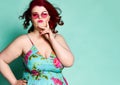 Choosy plus-size lady overweight woman in modern sunglasses and colorful sundress with her hand at her cheek chooses on mint Royalty Free Stock Photo