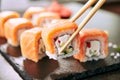 Chopstick holding Sushi Rolls Set with Salmon and Cream Cheese and Cuccumber on Black Slate Plate Close Up. Uramaki, Nori Maki or