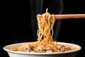 Chopsticks pick up tasty noodles with smoke on dark background. Ramen in white bowl.