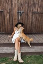 Chraming young blond woman in stylish cowboy boots, white dress and black hat, posing in front of barn wood wall Royalty Free Stock Photo