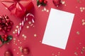 Christmas background with blank paper, red gift box with ribbon bow on red background and golden sparkling conffeti, candy cane,
