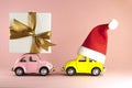 Christmas background. Little retro toy model car with Santa Claus hat and pink model car with present gift box on pastel