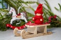 Christmas card. Bright red toy kid in knitted clothes on Santa`s sleigh on the background of a Christmas tree and a toy horse
